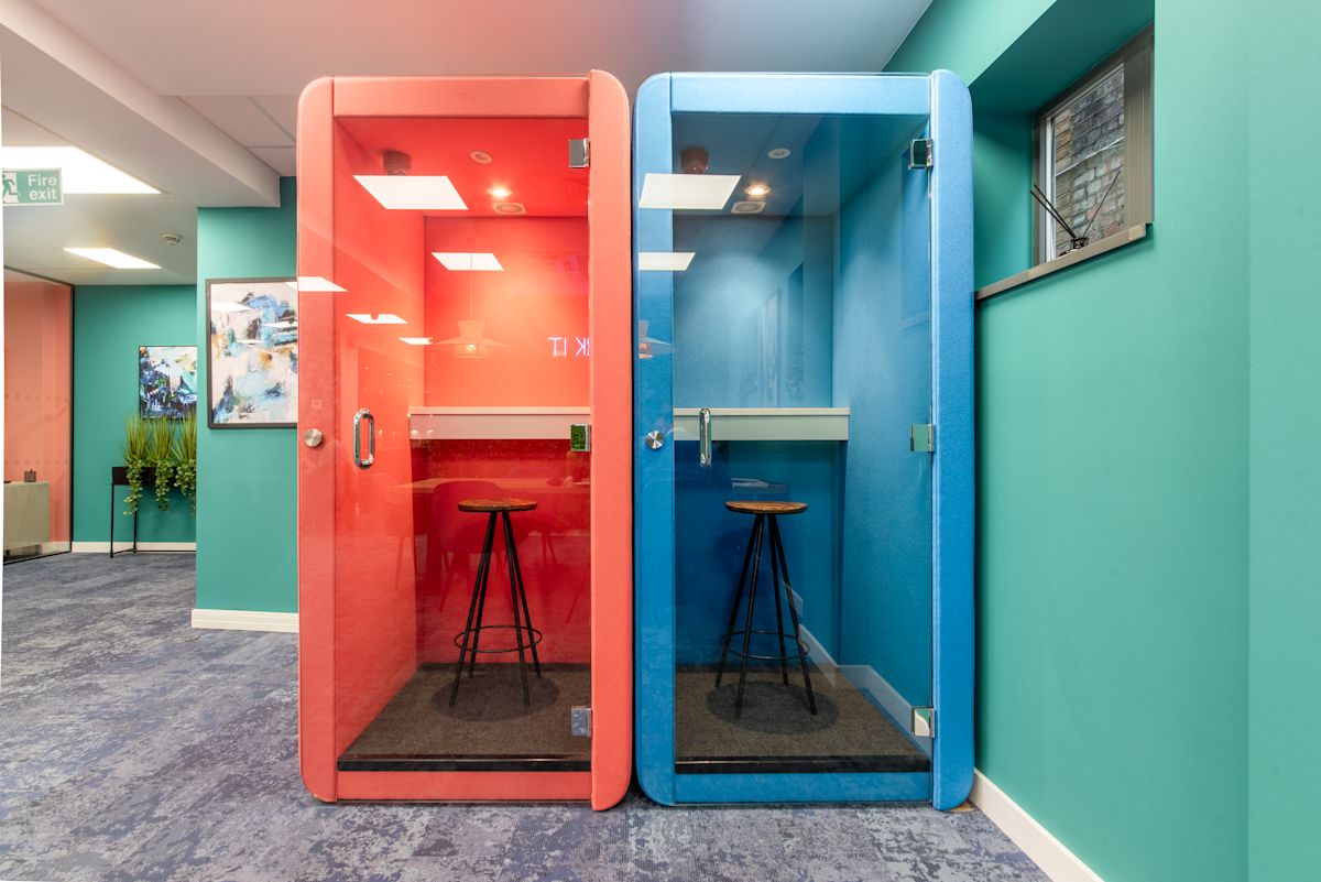 Two colorful phone booths, one red and one blue, with stools inside in a modern office setting.