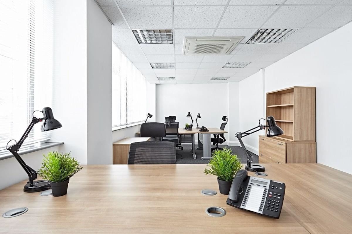 A modern, well-lit office space with wooden desks, black office chairs, desk lamps, and potted plants.