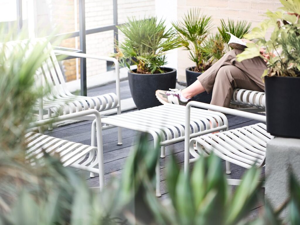 A person relaxes with a book on a patio with white metal chairs and potted plants on Wardour Street, London.