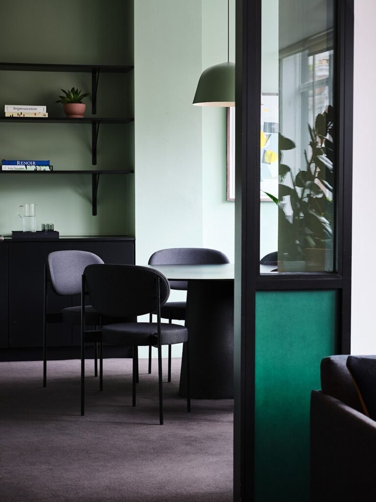 A modern office space with green walls, a round table, dark chairs, and shelves with books and a plant.