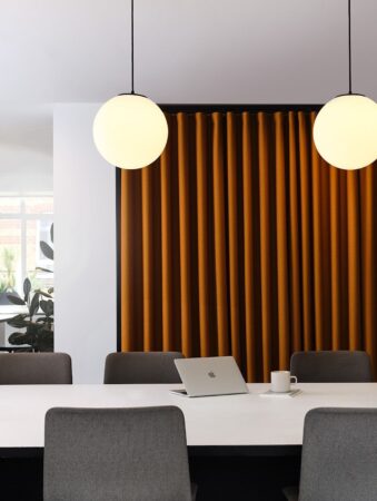 A modern conference room with a laptop and coffee cup on a white table, flanked by gray chairs and illuminated by spherical pendant lights.