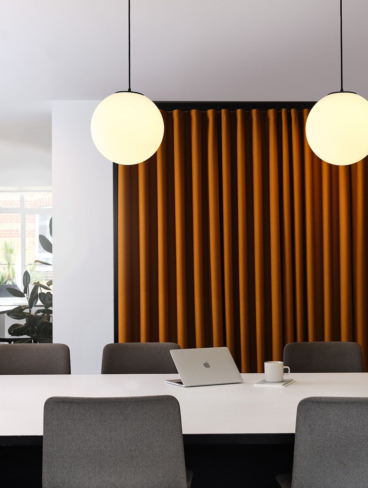 A modern conference room with a laptop and coffee cup on a white table, flanked by gray chairs and illuminated by spherical pendant lights.