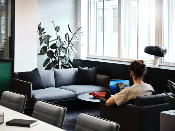 A person working on a laptop in a modern office lounge with a gray sofa and large windows.