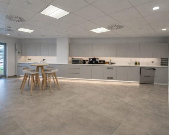 A modern office kitchen with a round table and stools, featuring gray cabinets and appliances along the wall.